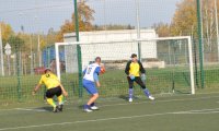 Zawodnik z żółtej koszulce i zawodnik w biało-niebieskiej koszulce podczas walki o piłkę 6 metrów od bramki, bramkarz na bramce. Piłka zmierzająca w światło bramki.