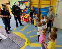 Policjant ubrany w kamizelkę przeciwuderzeniową i ochraniacze rąk i policjantka prezentują zaciekawionym dzieciom i opiekunkom wyposażenie funkcjonariusza oddziału prewencji.